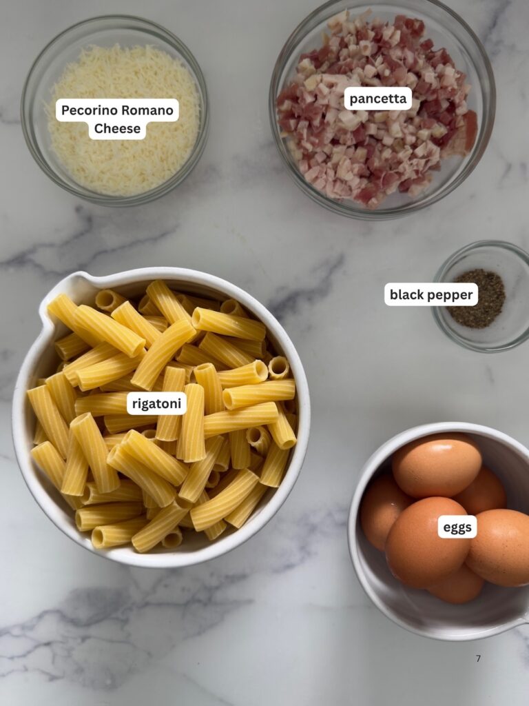rigatoni carbonara ingredients including rigatoni pasta, pancetta, eggs, Pecorino Romano cheese, and black pepper on a marble surface