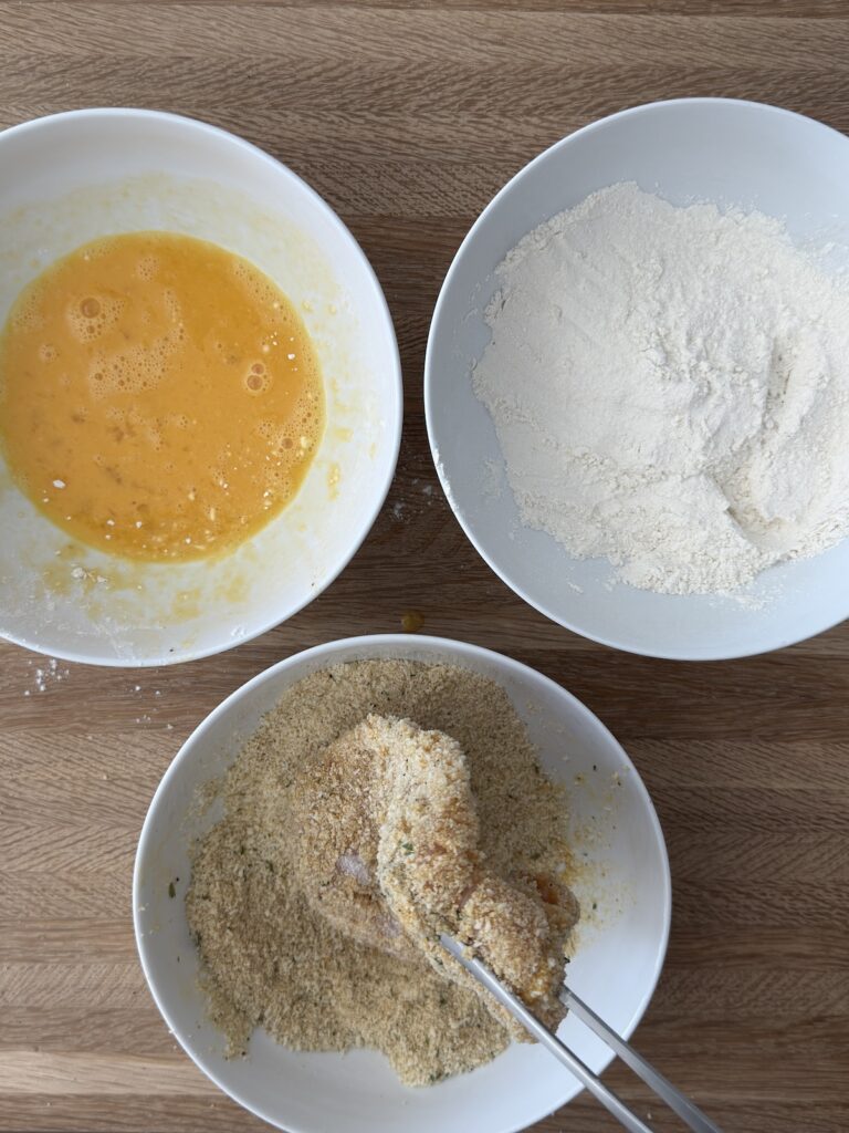Italian chicken cutlet being coated in seasoned breadcrumbs with bowls of egg wash and flour for breading station preparation.