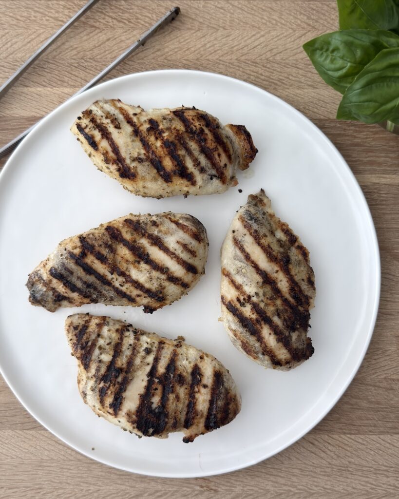 Grilled chicken breasts for Chicken Margherita on a white plate with char marks, photographed overhead on a wooden surface with fresh basil leaves