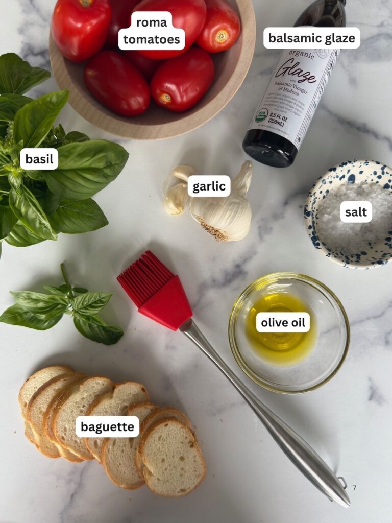 Ingredients for Bruschetta al Pomodoro including Roma tomatoes, fresh basil, garlic, extra virgin olive oil, flaky sea salt, balsamic glaze, and sliced baguette on a marble countertop.