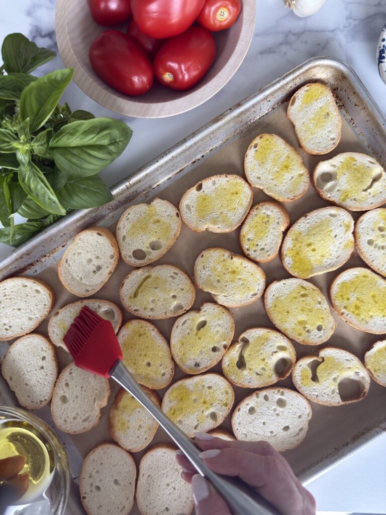Brushing olive oil onto sliced baguette bread on a baking sheet with fresh basil and Roma tomatoes for Bruschetta al Pomodoro.