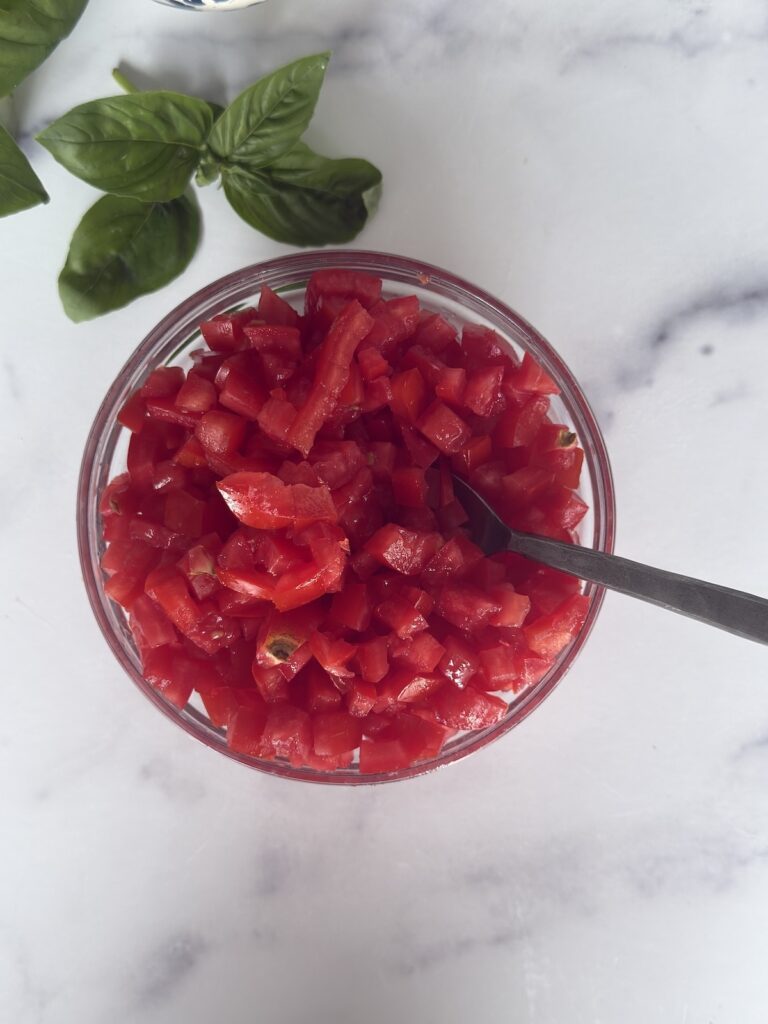 Bowl of diced Roma tomatoes for Bruschetta al Pomodoro with fresh basil on a marble countertop.