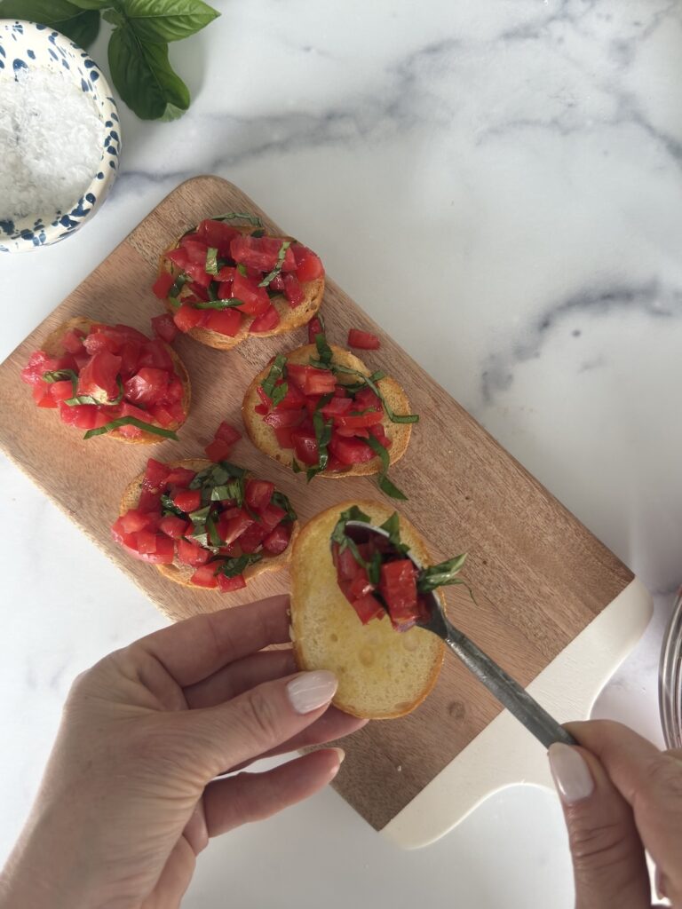 Hands topping toasted baguette slices with diced tomatoes and fresh basil for Bruschetta al Pomodoro on a wooden serving board.