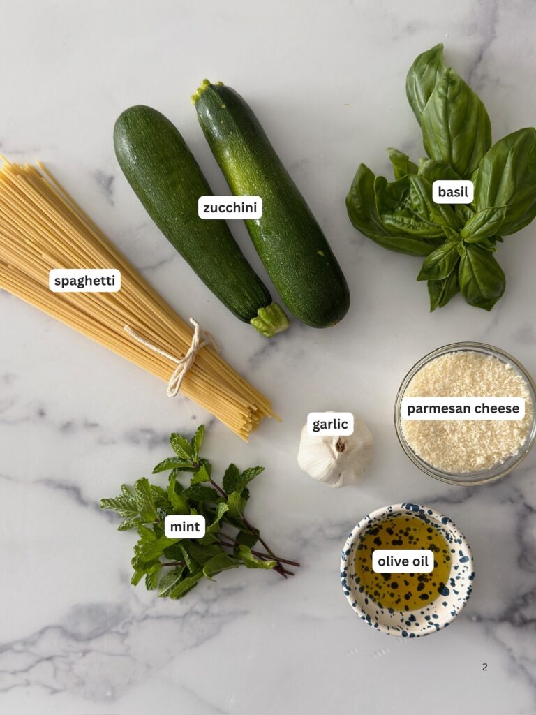 Zucchini pasta ingredients including spaghetti, fresh zucchini, basil, mint, garlic, olive oil, and Parmesan cheese, arranged on a marble surface.