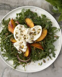 Grilled peach burrata salad with arugula, red onion, toasted pistachios, and creamy burrata on a white platter.