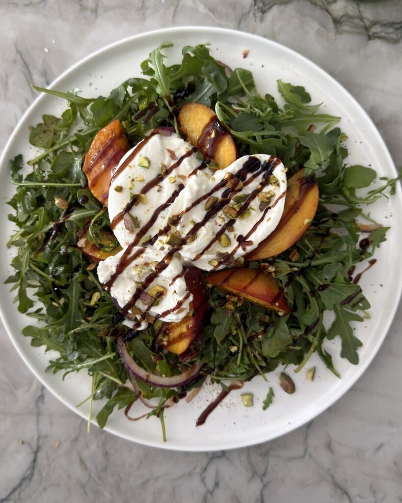 Grilled peach burrata salad with balsamic glaze, creamy burrata, arugula, red onion, and toasted pistachios.