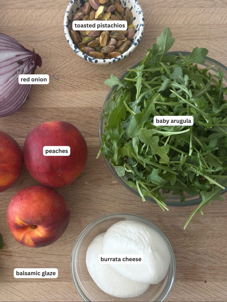 Ingredients for a peach and burrata salad including fresh peaches, creamy burrata cheese, baby arugula, roasted pistachios, red onion, and balsamic glaze, arranged on a wooden board.