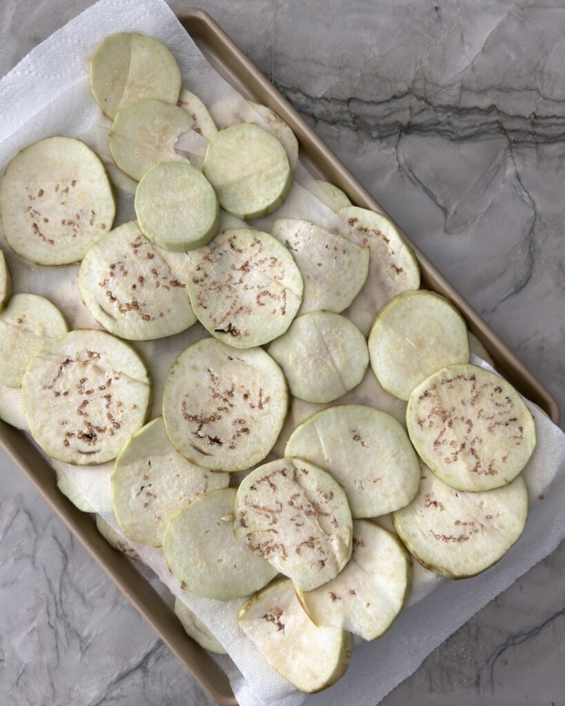 Peeled eggplant slices sprinkled with salt and resting on a paper towel-lined baking sheet to draw out excess moisture.
