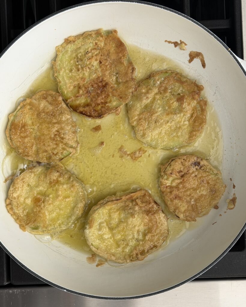 Flour- and egg-coated eggplant slices frying until golden in olive oil in a white skillet.