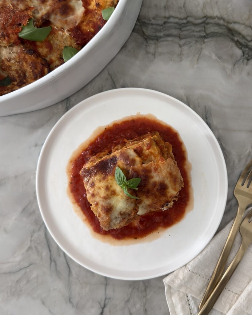 A serving of eggplant Parmesan with golden melted mozzarella and fresh basil over marinara sauce on a white plate.