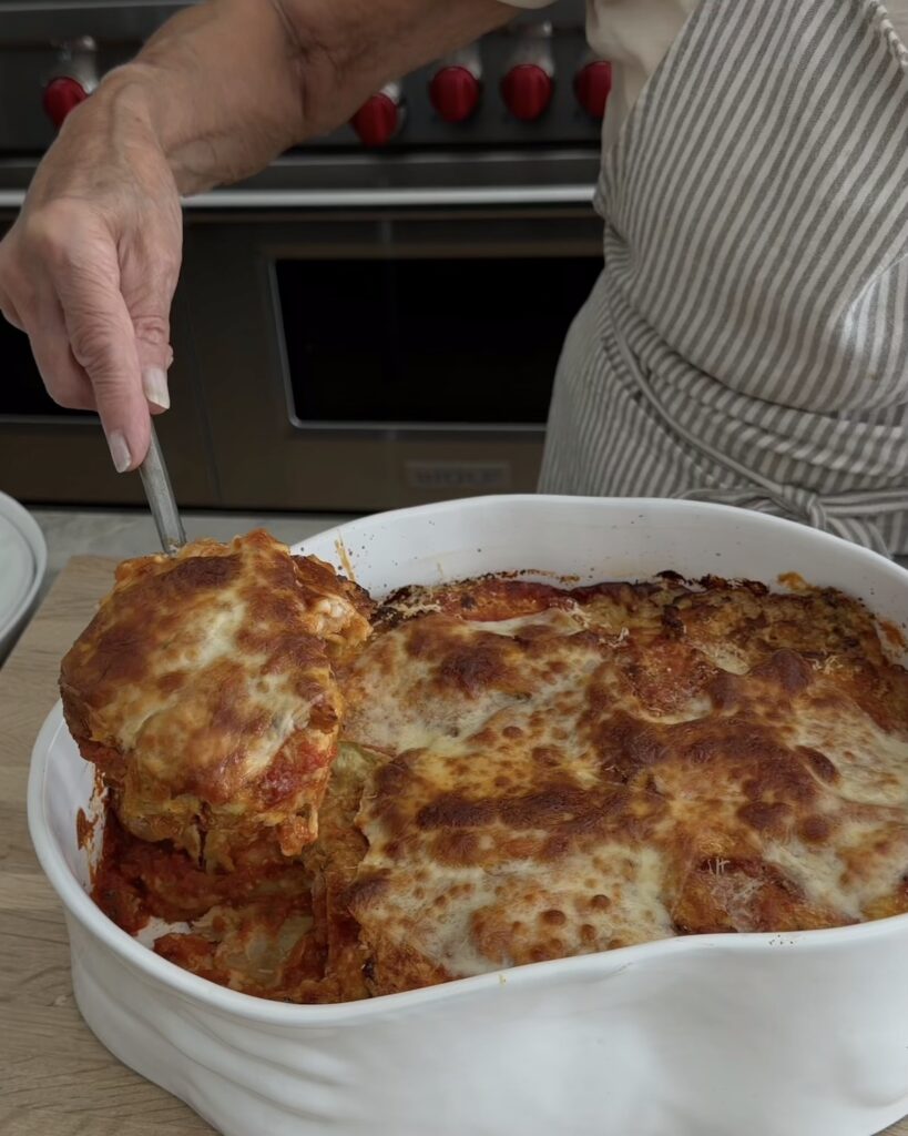 A slice of eggplant Parmesan lifted from a white baking dish, showing layers of eggplant, marinara sauce, and golden-brown melted mozzarella.