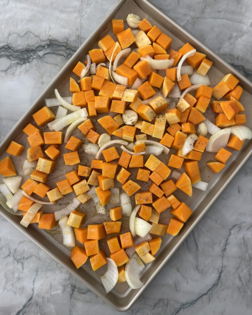 Uncooked butternut squash cubes, onion wedges, and garlic spread on a rimmed baking sheet before roasting.