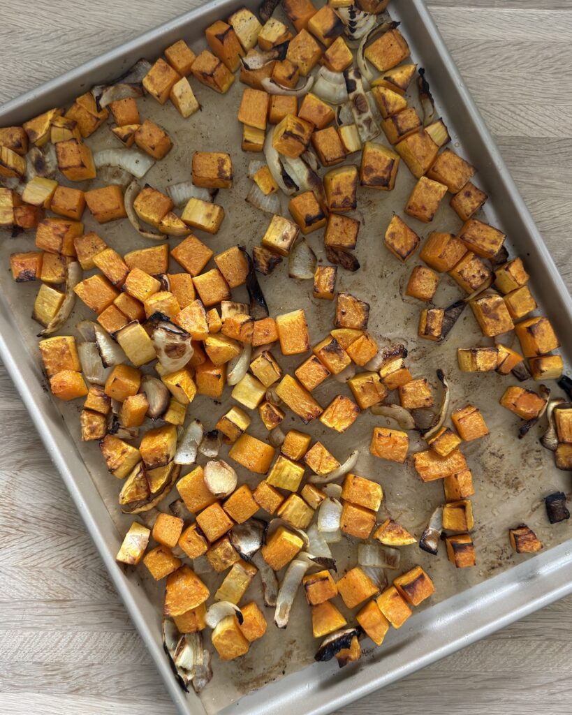 Roasted butternut squash cubes, onion, and garlic with browned edges on a rimmed baking sheet.