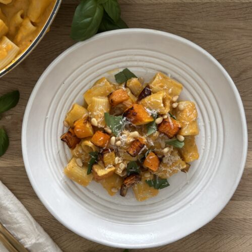 Creamy butternut squash pasta topped with roasted squash cubes, Parmesan, basil, and pine nuts in a white bowl.