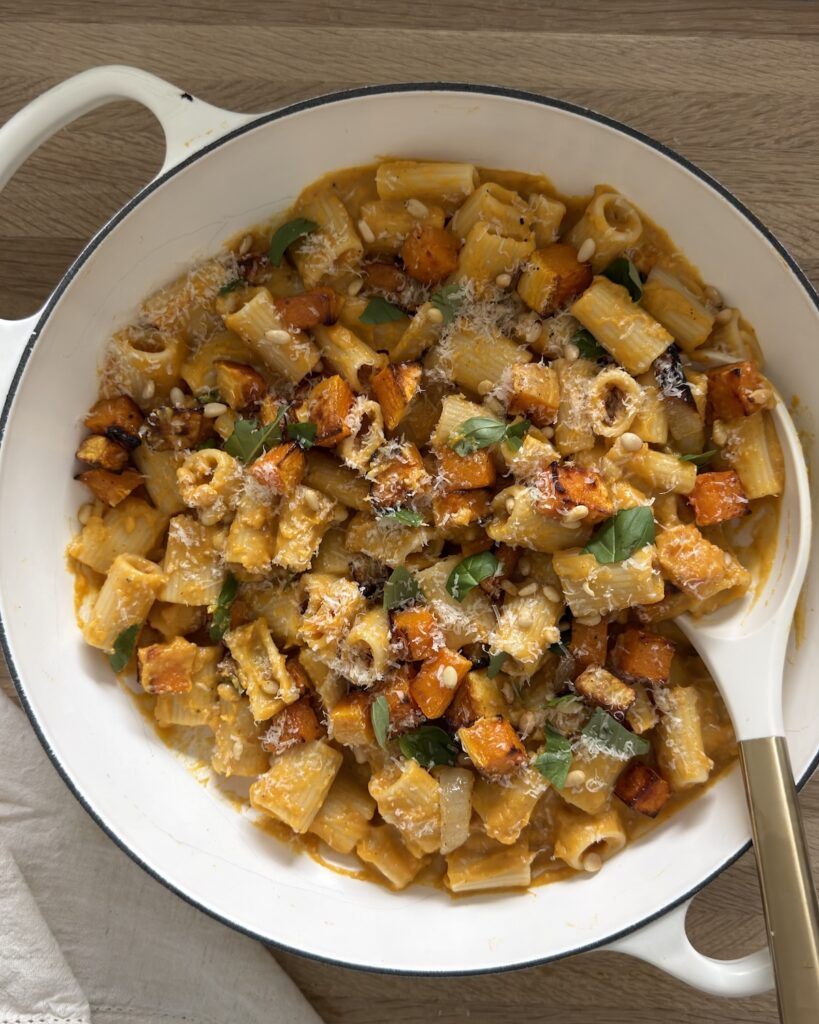 Rigatoni in creamy butternut squash sauce with roasted squash cubes, Parmesan, basil, and pine nuts in a white skillet.