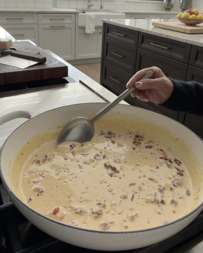 Creamy sun-dried tomato sauce being stirred in a white skillet before adding the chicken, spinach, and pasta.