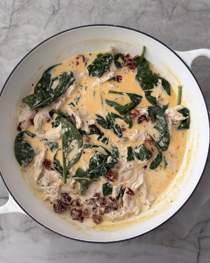 Shredded chicken, spinach, and sun-dried tomatoes in a creamy sauce in a white skillet before adding the pasta.