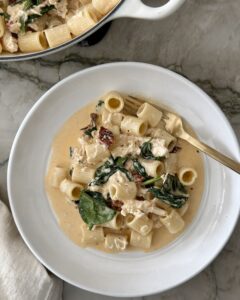 Creamy chicken sun-dried tomato pasta with rigatoni and spinach in a white bowl, with a gold fork resting on the rim.