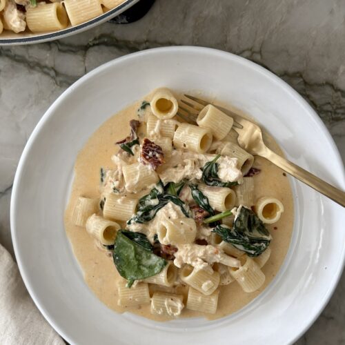 Creamy chicken sun-dried tomato pasta with rigatoni and spinach in a white bowl, with a gold fork resting on the rim.