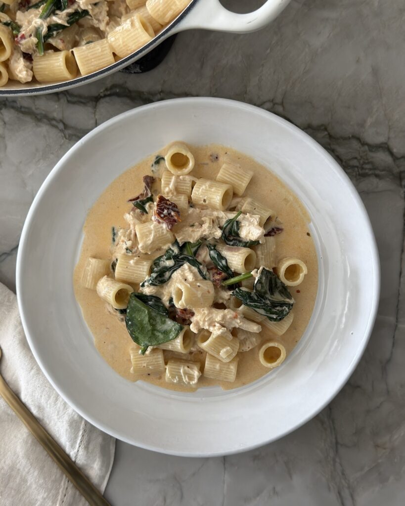 Overhead view of creamy chicken sun-dried tomato pasta with rigatoni and spinach in a white bowl.