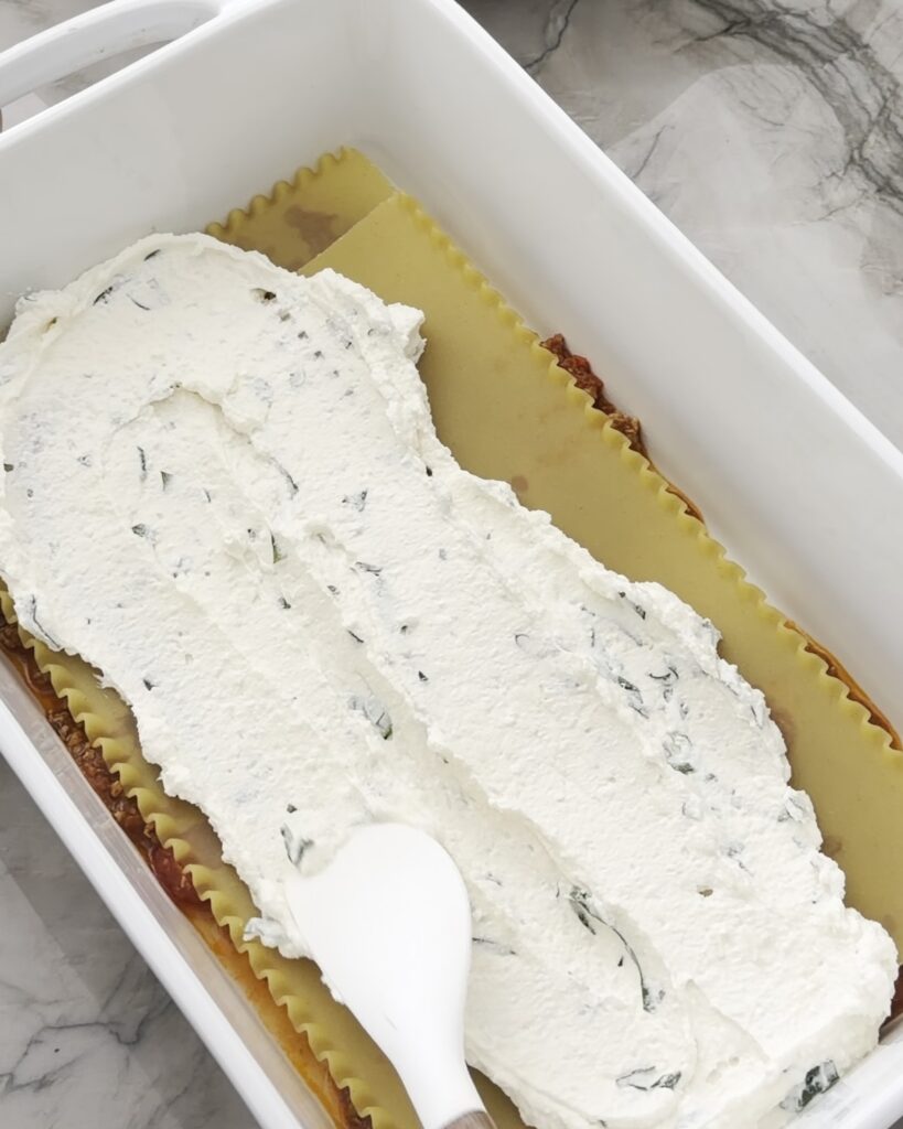 Ricotta filling is being spread over lasagna noodles in a white baking dish.