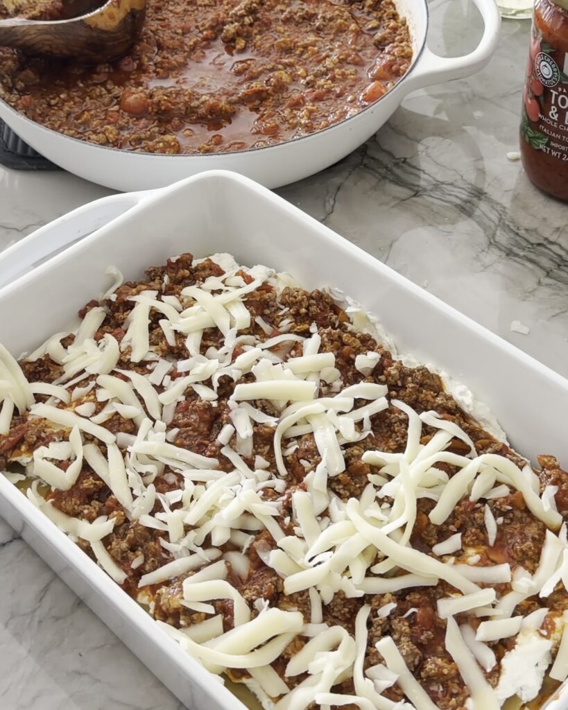 Shredded mozzarella scattered over a layer of meat sauce while assembling homemade lasagna in a white baking dish.