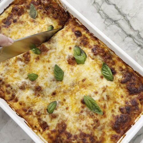 Easy homemade lasagna topped with golden-brown melted cheese and fresh basil, being sliced in a white baking dish.