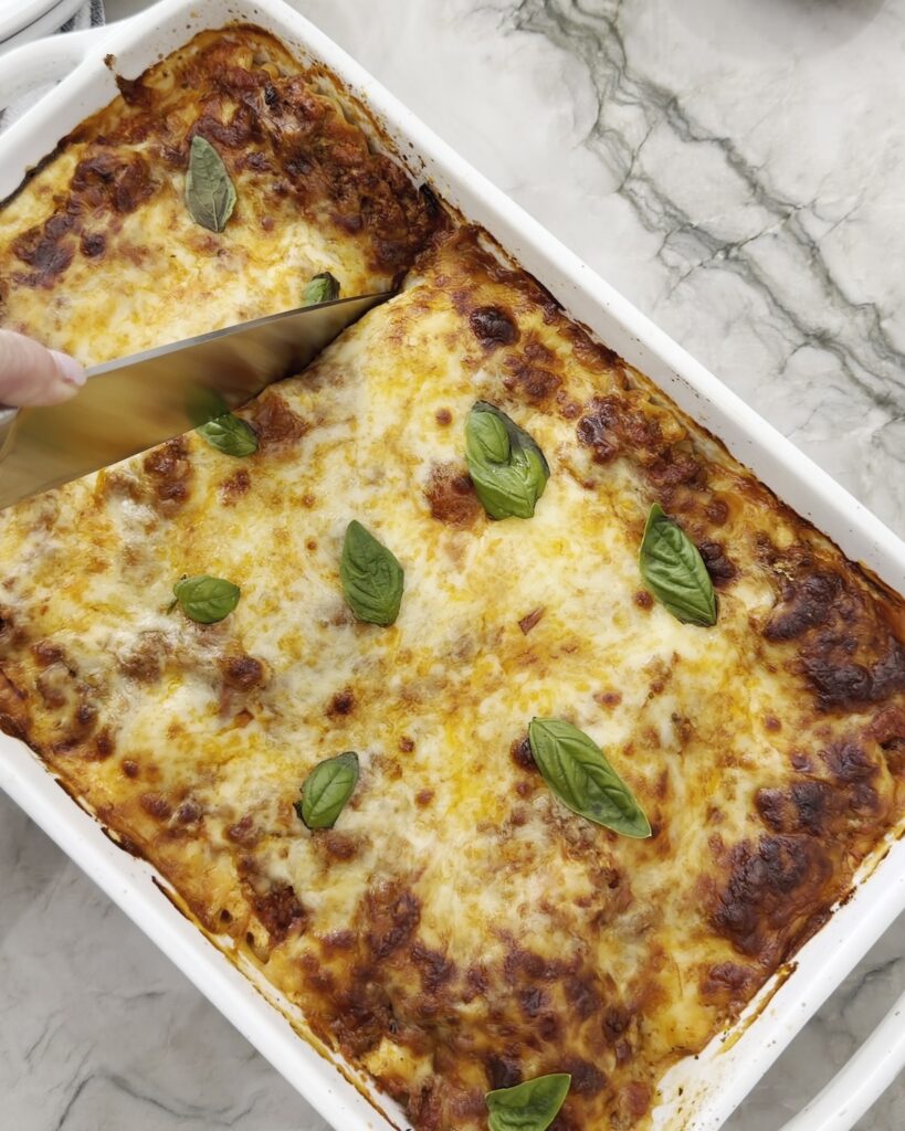 Easy homemade lasagna topped with golden-brown melted cheese and fresh basil, being sliced in a white baking dish.