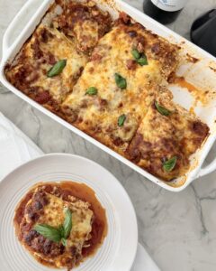 Homemade lasagna with browned cheese and fresh basil in a white baking dish, with a slice served on a plate beside it.