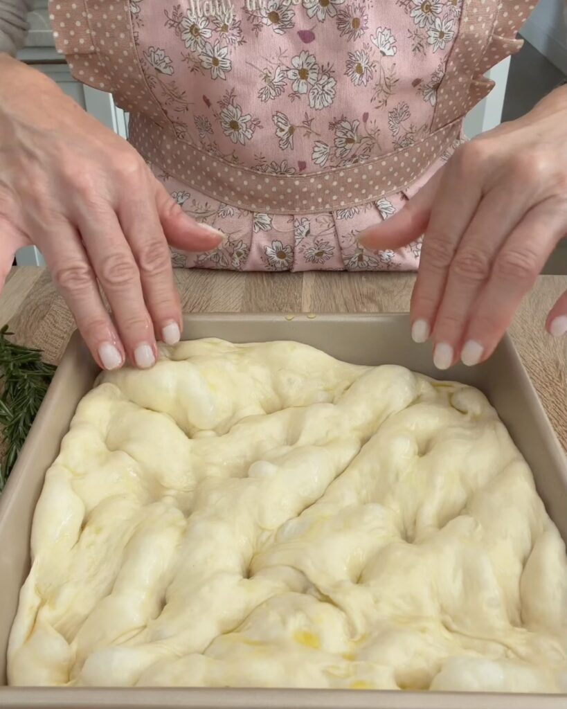 Hands dimpling pizza dough coated with olive oil in a square metal baking pan.