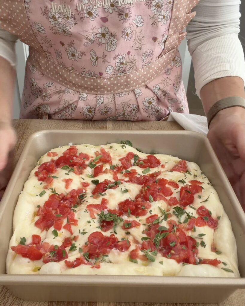 Dimpled pizza dough topped with chopped cherry tomatoes and fresh oregano in a square baking pan before baking.