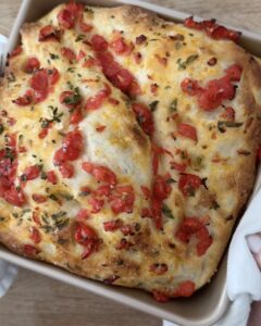Golden pizza dough focaccia topped with chopped cherry tomatoes and fresh oregano in a square baking pan.