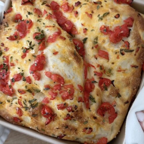 Golden pizza dough focaccia topped with chopped cherry tomatoes and fresh oregano in a square baking pan.