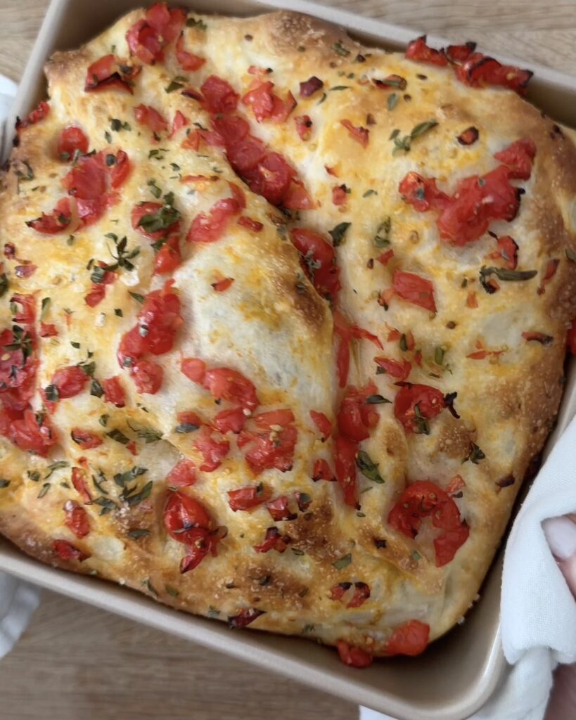 Golden pizza dough focaccia topped with chopped cherry tomatoes and fresh oregano in a square baking pan.