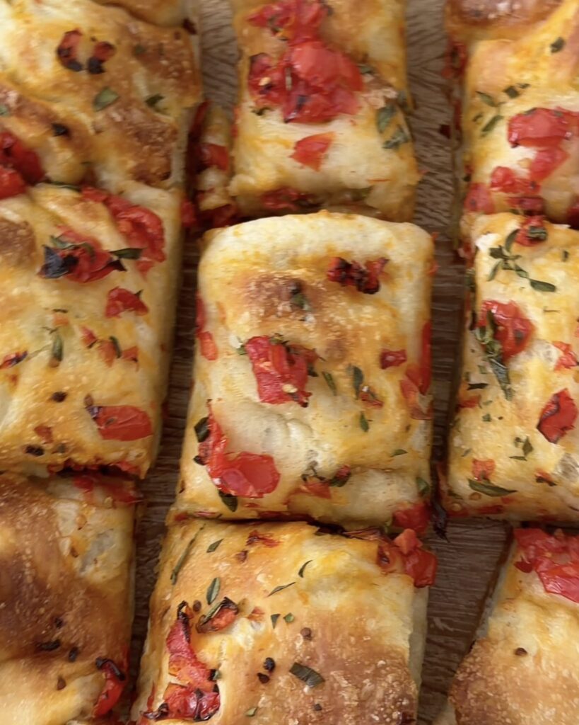 Pizza dough focaccia topped with cherry tomatoes and fresh oregano, cut into squares on a wooden board.